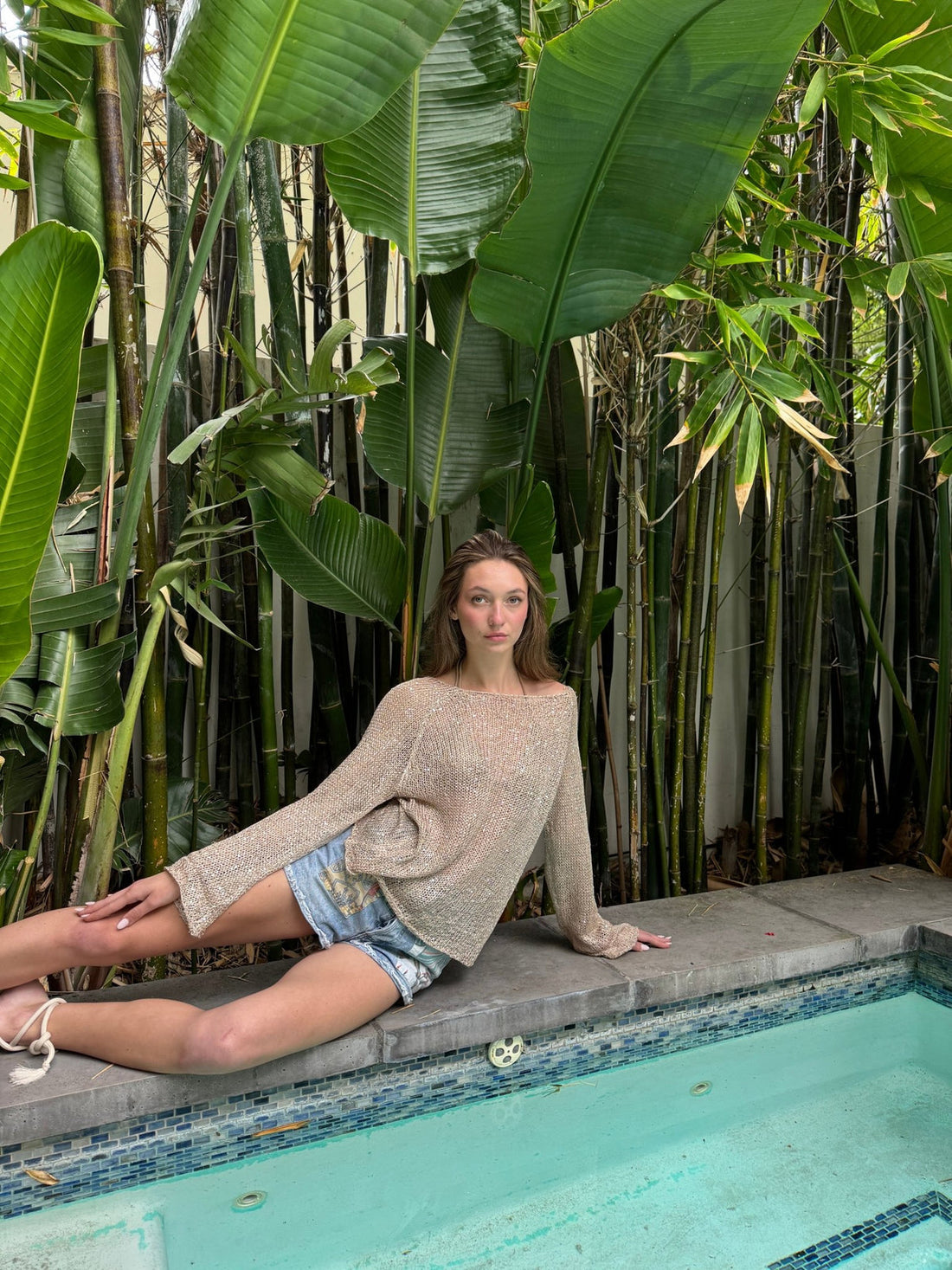 Woman sitting by a pool among bamboo and banana leaves in a beige open-knit sweater and denim shorts
